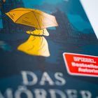 Text: "SPIEGEL Bestseller Autorin", "DAS MÖRDER". Illustration: Person im gelben Mantel mit Regenschirm vor dunkler Kulisse.