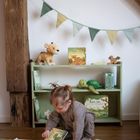 Ein kleines Kind liest Bücher vor einem Regal. Darauf Kuscheltiere, Kinderbücher und eine Girlande.
