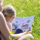 Zwei Personen lesen gemeinsam ein buntes Kinderbuch mit Tierillustrationen auf einer Blumenwiese.
