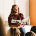 Ein Mann mit langem Haar liest einer Gruppe von Kindern aus einem Buch vor. Er sitzt auf einem Tisch in einem hellen Raum.