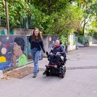 Zwei Personen auf einem Gehweg, eine Frau führt einen Rollstuhlfahrer. Bunte Wandmalerei mit Kindern und Planetensystem.