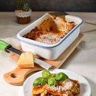 Eine Lasagne in einer Auflaufform mit einem Stück auf einem Teller daneben, garniert mit Basilikum und geriebenem Käse.
