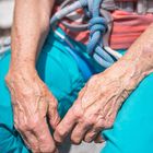Zwei ältere Hände mit sichtbaren Adern, ein Klettergurt um die Taille, bunte Kleidung in Rot- und Blautönen.