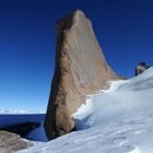 Ein imposanter, steil aufragender Felsen erhebt sich aus einer schneebedeckten Landschaft unter einem klaren, tiefblauen Himmel.
