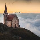 Eine kleine Steinkapelle mit rotem Dach auf einem Berggipfel, umgeben von Wolken im Sonnenaufgang.