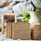 Mehrere Scheiben Brot vor einem Laib, mit rustikalem Hintergrund und einem Krug im unscharfen Hintergrund.