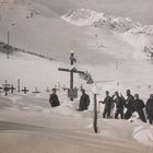 Mehrere Gräber mit Holzkreuzen auf einem verschneiten Feld; Menschen stehen dahinter vor schneebedeckten Bergen.