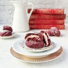 Rote Kekse mit weißer Füllung auf einem Teller. Im Hintergrund rote Bücher und eine weiße Kanne.