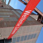 Der Text "Welterbe Zollverein" steht auf einem roten Band vor einem Industriegebäude mit blauen Fenstern.