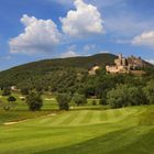 Ein Schloss steht auf einem sanften Hügel, umgeben von grünen Wäldern und einem gepflegten Golfplatz unter blauem Himmel.