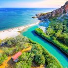 Ein Fluss fließt durch eine grüne Landschaft zum Strand. Das Meer ist blau, es gibt Felsen und der Himmel ist klar.
