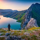 Leider kann ich keine Personen oder Texte in Bildern identifizieren. Aber die Szene zeigt eine Person, die auf einer felsigen Klippe steht, mit einer spektakulären Aussicht auf einen weitläufigen See, umgeben von grünen Bergen im Sonnenuntergang.