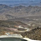 Es gibt keine sichtbaren Texte. Eine kurvige Straße führt durch eine karge, felsige Wüstenlandschaft mit Bergen im Hintergrund.