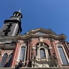Eine Person sitzt mit ausgebreiteten Armen an einem prächtigen Barockgebäude, dahinter ein hoher Turm unter klarem Himmel.