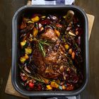 Kein Text. Ein gebratener Braten mit Rosmarin in einer dunklen Auflaufform, umgeben von buntem Gemüse und einem Tuch darunter.