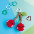 Zwei gehäkelte Kirschen mit lächelnden Gesichtern an einem Ring auf blau-grünem Hintergrund, umgeben von Herzformen.