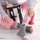 Keine Texte zu sehen. Eine Person trägt rosa Hosen und schwarz-weiße Socken mit Pinguinmustern. Ein schwarzer Stoffbeutel mit Wolle steht daneben.