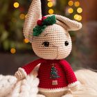 Ein gehäkelter Hase mit Weihnachtsbaum auf rotem Pullover, trägt ein grünes Blatt und rote Beeren auf dem Kopf, vor Lichtern.