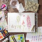 Illustration von Blumen im Skizzenbuch, umgeben von Wasserfarben, Pinseln und Mustern auf einem Holztisch.