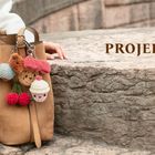 Text: "PROJEKTE". Ein hellbrauner Rucksack mit bunten, gehäkelten Anhängern auf einem Stein, daneben eine Hand.