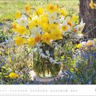 Text: "MÄRZ". Gelbe und weiße Narzissen in einer Vase, umgeben von Frühjahrsblumen auf einer Wiese. Rechts ein Korb mit Eiern.