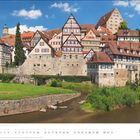 Text: "MÄRZ". Häuser mit Fachwerk an einem steinernen Flussufer, grüne Wiese, blauer Himmel mit Wolken.