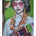 Text: "03" unten links. Darstellung (vermutlich Graffiti): Frau mit buntem Kopfschmuck und großen, farbigen Halsketten. Grüne Wand.