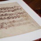 Ein Notenblatt in einem geöffneten Buch, auf einem Holztisch liegend, zeigt handgeschriebene Musiknoten.