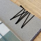 Schwarze Fäden durchlöchern graue Pappe und formen das Muster "WW". Darunter blau-weiß kariertes Band auf Holzuntergrund.