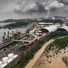 Industrielandschaft mit Hafen, Kränen, großer Struktur mit Kuppeldächern, rauchenden Schornsteinen und Umgebung mit Vegetation.