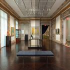 Galerie mit kunstvoll verzierten Wänden, Gemälden, Glaskabinett, dunkelbraunem Boden und Bank. Exit-Schilder über Türen.