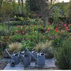 Kalender: Mai. Bunt blühender Garten vor einem Holzhaus mit roten Dachziegeln. Sitzbank mit Blumentöpfen.