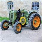 Auf einem Hof steht ein grüner, antiker Traktor. Links steht senkrecht "Lanz Aul". Die Räder sind groß und gelb-orange.