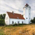 Ein weißes Haus mit rotem Ziegeldach steht neben einem weißen Leuchtturm. Im Vordergrund ist trockene Wiese.