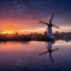 Sonnenuntergang reflektiert auf einem ruhigen Fluss, silberne Windmühle am Ufer, rötliche Wolken im Himmel.