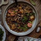 Ein Teller mit Linseneintopf, gefüllt mit Karotten, Kartoffeln, Pilzen und Kräutern, liegt auf einem Holztisch mit Besteck.