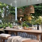 Ein gedeckter Holztisch im Freien, umgeben von Pflanzen, mit Kerzen, Gläsern und einer gestrickten Decke auf der Bank.