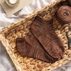 Auf einem Korb aus geflochtenem Material liegen gestrickte Socken. Daneben sind Wolle, Schere und eine Kerze.