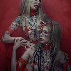 Zwei Personen in roten, stachelbesetzten Outfits mit silbernen Ketten vor rotem Hintergrund, dramatisches Make-up und Tattoos.