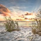 Sonnenuntergang über dem Meer, sandiger Strand mit Dünen und Grasbüscheln, ruhige Wellen, orange-lila Wolken am Himmel.
