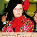 "Ich umarmte die Kunst als Erlösung und Notwendigkeit." Frau mit rotem Schal und bunter Kleidung, schwarzer Hut mit Blume.