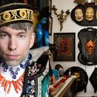 Links ein Mann in aufwendiger Kleidung, rechts eine Wand mit Masken und Bilderrahmen, darunter asiatische Motive.