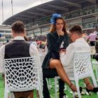 Logos: Mehrmals "Investec". Frau mit blauem Hut und Make-up sitzend, umgeben von eleganten Menschen und Flaschen.