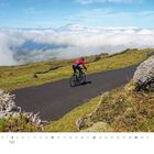 Text "März". Ein Radfahrer in roter Jacke fährt auf einem kurvigen Weg durch eine grüne Hügellandschaft.