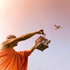 Ein Mensch in orangefarbener Robe lässt Vögel aus einem kleinen hölzernen Käfig fliegen, vor hellem Himmel.