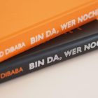 "BIN DA, WER NOCH?" auf Rücken von zwei Bücher, eines orange, eines schwarz.