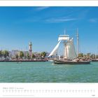 März 2027 | Am Meer. Ein Segelschiff segelt vor einer Hafenlandschaft mit einem Leuchtturm bei klarem Himmel.