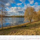 Kalender mit März 2027. Bild zeigt einen ruhigen See, umgeben von Bäumen unter blauem Himmel mit Wolken.