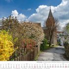 Kalendertext: März 2027. Hückeswagen – Am Kamp mit Blick zur Kirche. Blühende Sträucher und ein Kirchturm vor blauem Himmel.