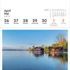 Kalender mit Monatsübersicht für April und Mai 2027. Darunter ein See mit zwei Bootshäusern und Bäumen bei klarem Himmel.